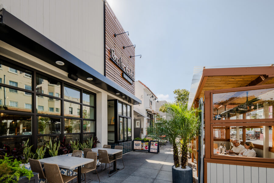 Outdoor seating area at a modern restaurant with tables, chairs, potted plants, and a wooden dining enclosure where people are eating.