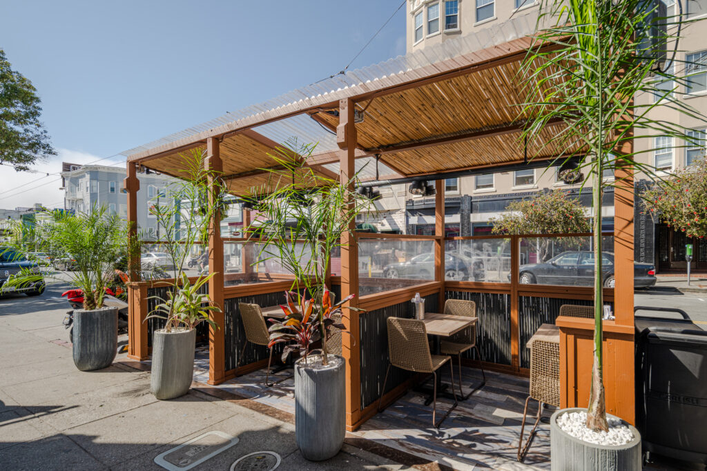 Outdoor dining area with wooden pergola, wicker chairs, and tables, separated by clear panels; potted plants line the structure on a city sidewalk under clear skies.