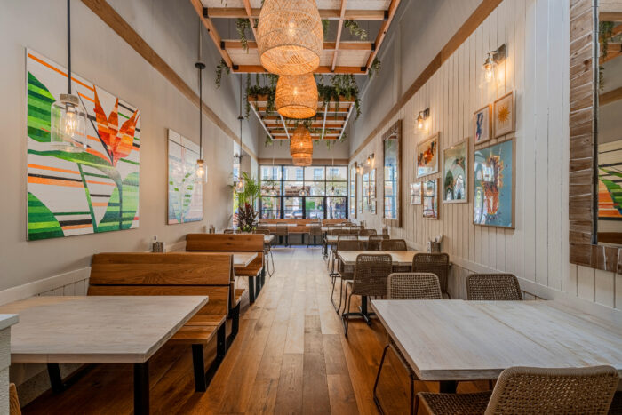 A modern, well-lit restaurant interior with wooden floors, booth and table seating, woven light fixtures, and colorful artwork on white walls.