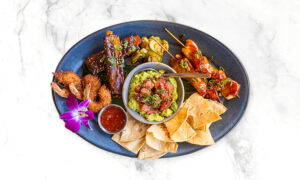 A blue plate with grilled ribs, coconut shrimp, chips, poke, grilled skewers, guacamole, dipping sauce, and a purple flower on a white marble surface.