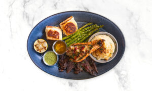 A blue oval plate with grilled asparagus, mashed potatoes, sliced steak, a grilled lobster tail, two rolls, grilled lemon halves, and two small cups of sauce.