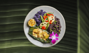 A plate with assorted vegetables, black rice, a grilled lemon wedge, a banana leaf-wrapped item, and a purple orchid garnish on a green leaf background.