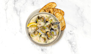 A bowl of clam chowder garnished with lemon and green onions, served with two slices of toasted bread on a marble surface.
