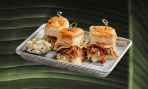 A metal tray holding three pulled pork sliders with coleslaw on buns, each secured with a pick, and a side of macaroni salad. The tray is placed on newspaper over a leaf background.