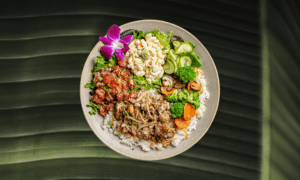A bowl of white rice topped with pulled pork, macaroni salad, mixed greens, poke, sliced cucumbers, broccoli, carrots, and an orchid flower garnish, placed on a green surface.