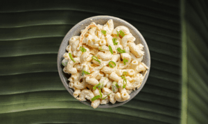 A bowl of macaroni salad garnished with chopped green onions, placed on a large green leaf background.