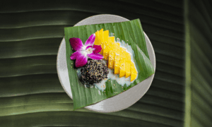 A plate with sliced mango, black sticky rice topped with sesame seeds, coconut milk, and a purple orchid, served on a banana leaf.