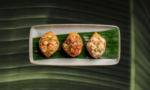 Three pieces of sushi with different toppings are arranged in a row on a rectangular plate lined with a green leaf, set on a leafy background.