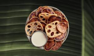 A bowl of lotus root and sweet potato chips with a small metal cup of dipping sauce, set on a green leaf background.