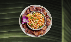 A round plate with purple sweet potato chips surrounding a bowl of diced poke with vegetables, a lemon wedge, and an orchid garnish, placed on a green leaf background.