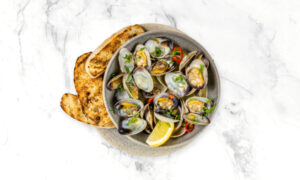 A plate of cooked clams garnished with herbs and sliced chili, served with lemon wedges and toasted bread on a marble surface.