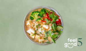 A bowl of soup with tofu, broccoli, red bell pepper, herbs, noodles, and a creamy broth, placed on a light green surface with the 
