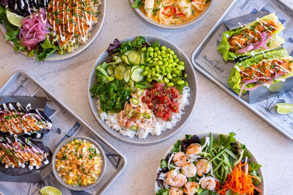 An overhead view of assorted poke bowls, shrimp salad, lettuce wraps, sushi rolls, and corn on a light table at a vibrant West Coast restaurant.